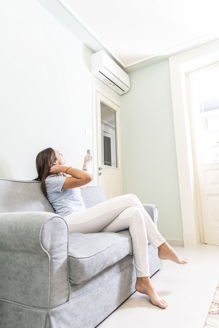 Mujer relajada sentada en sofá gris en una habitación luminosa, mirando un aire acondicionado instalado cerca del techo
